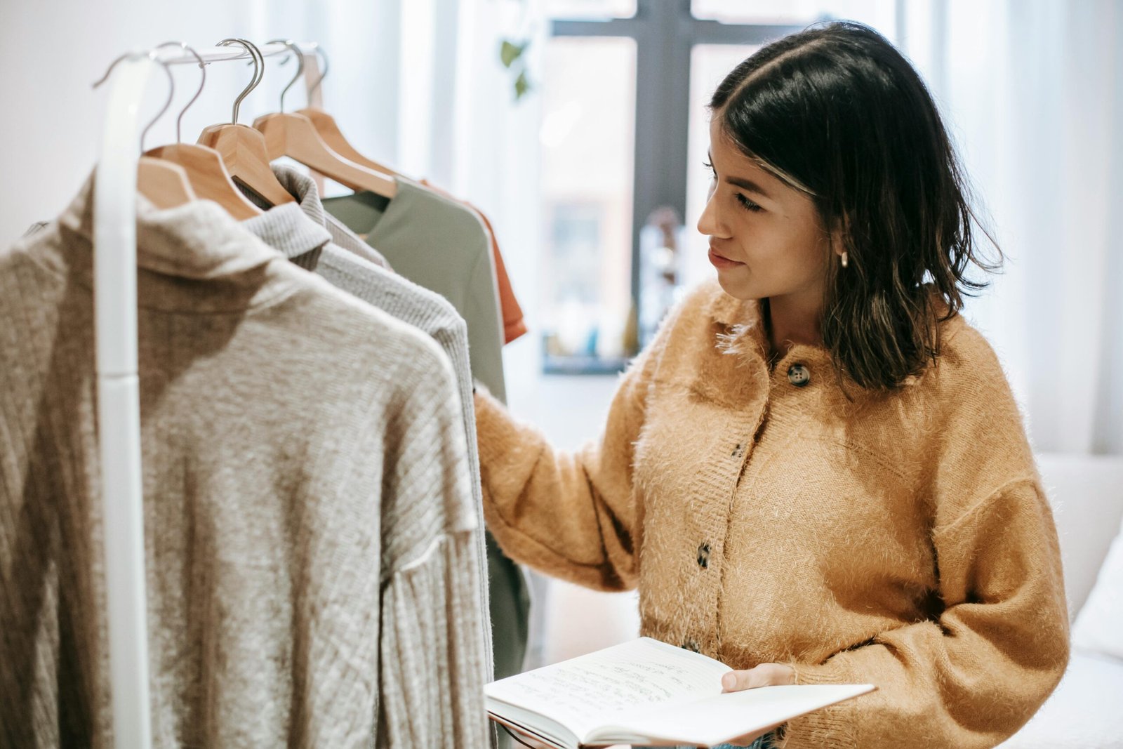 A woman designer examines clothing on a rack indoors, holding a notebook.